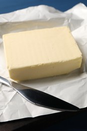 Block of tasty butter in open foil packaging and knife on blue wooden table, closeup