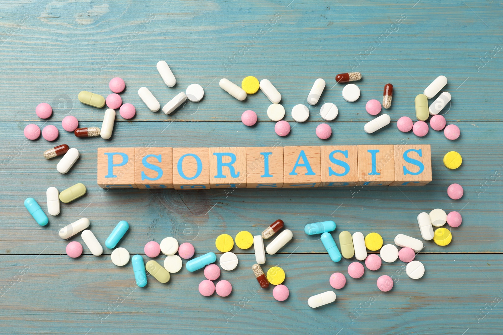 Photo of Word Psoriasis made of cubes with letters and pills on light blue wooden table, flat lay