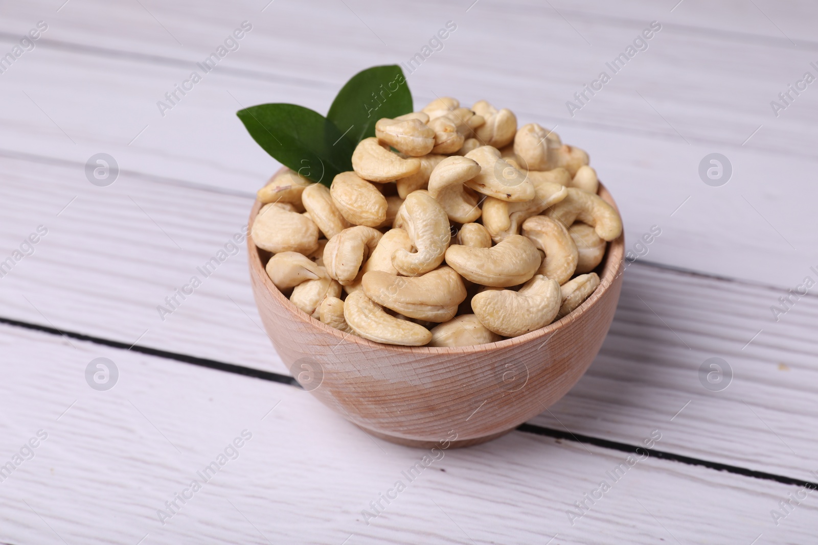 Photo of Tasty cashew nuts and green leaves in bowl on light wooden table, closeup