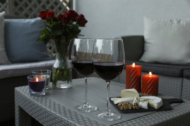 Glasses of wine, vase with roses, burning candles and snacks on rattan table at balcony in evening, closeup