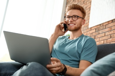 Young man talking on phone while using laptop in living room