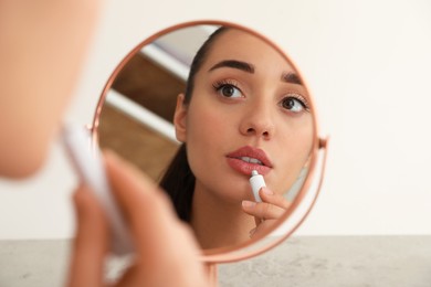 Woman with herpes applying cream on lips in front of mirror against light background