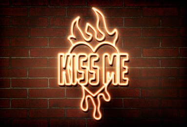 Glowing neon sign with words Kiss Me, heart and flames on brick wall