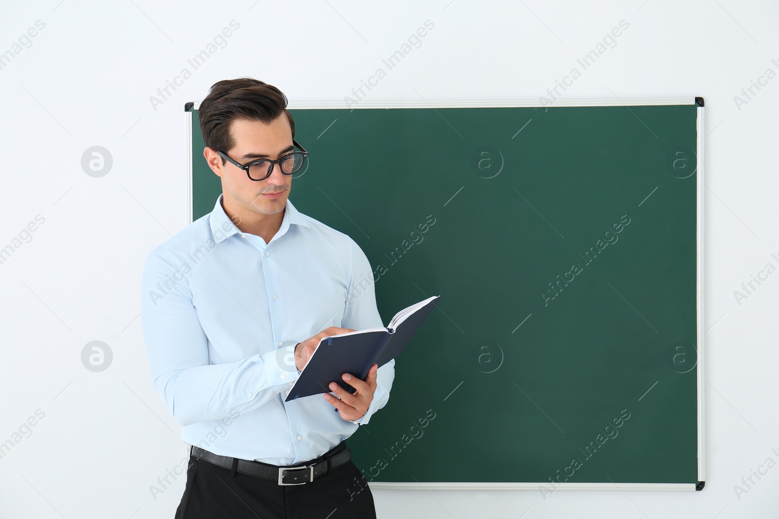 Photo of Young teacher with book near blank chalkboard in classroom. Space for text