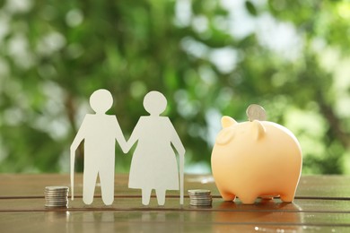 Pension savings. Figure of senior couple, piggy bank and stacked coins on wooden table outdoors