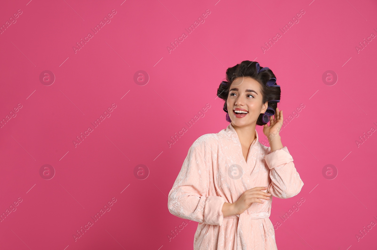 Photo of Happy young woman in bathrobe with hair curlers on pink background, space for text