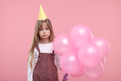 Photo of Bored little girl in party hat with bunch of balloons on pink background