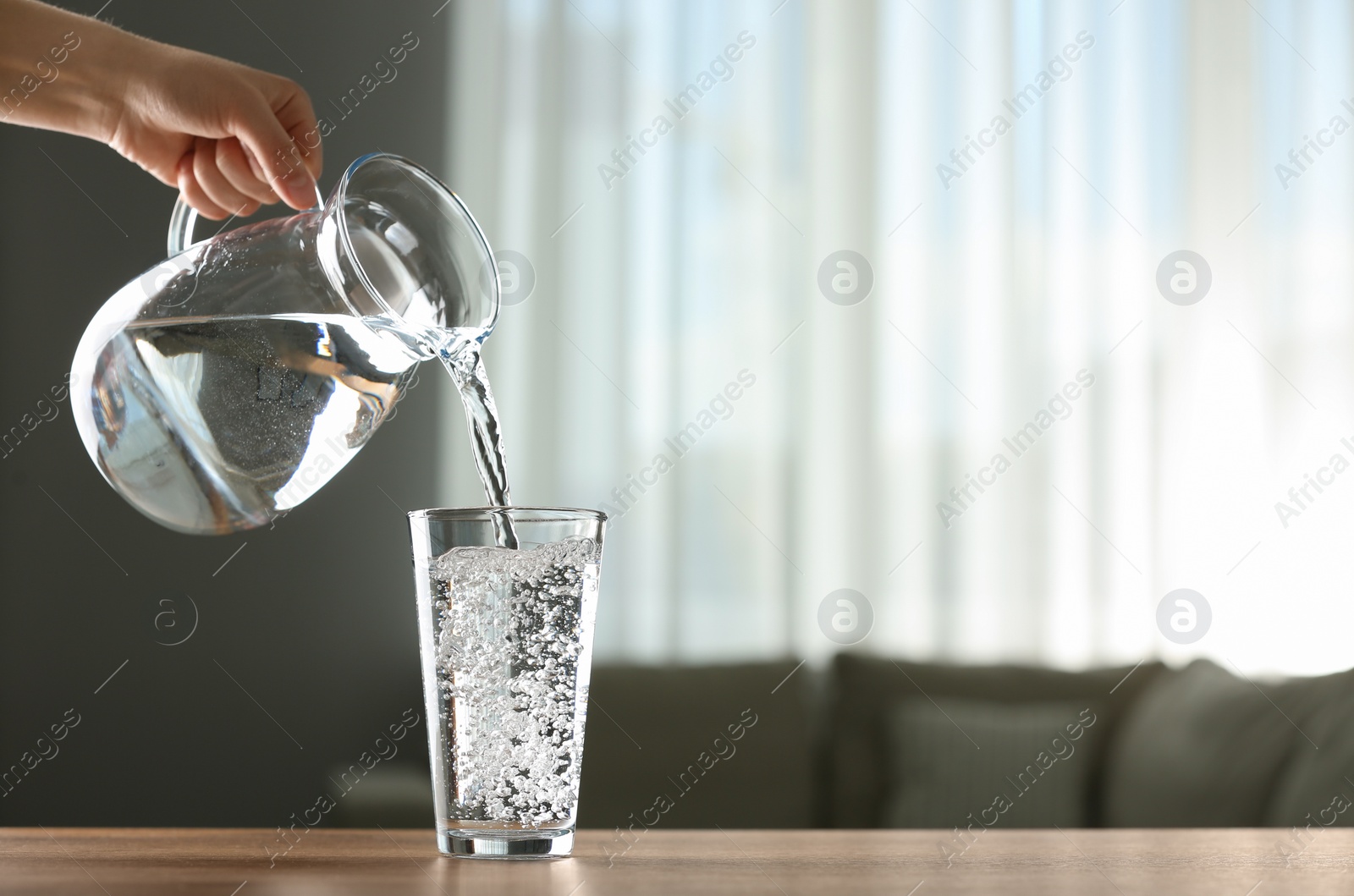 Photo of Woman pouring water from jug into glass at wooden table indoors, closeup. Space for text