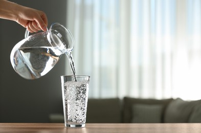 Woman pouring water from jug into glass at wooden table indoors, closeup. Space for text