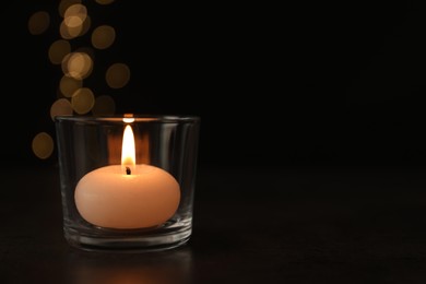 Photo of Burning candle in glass holder on table against blurred lights, space for text