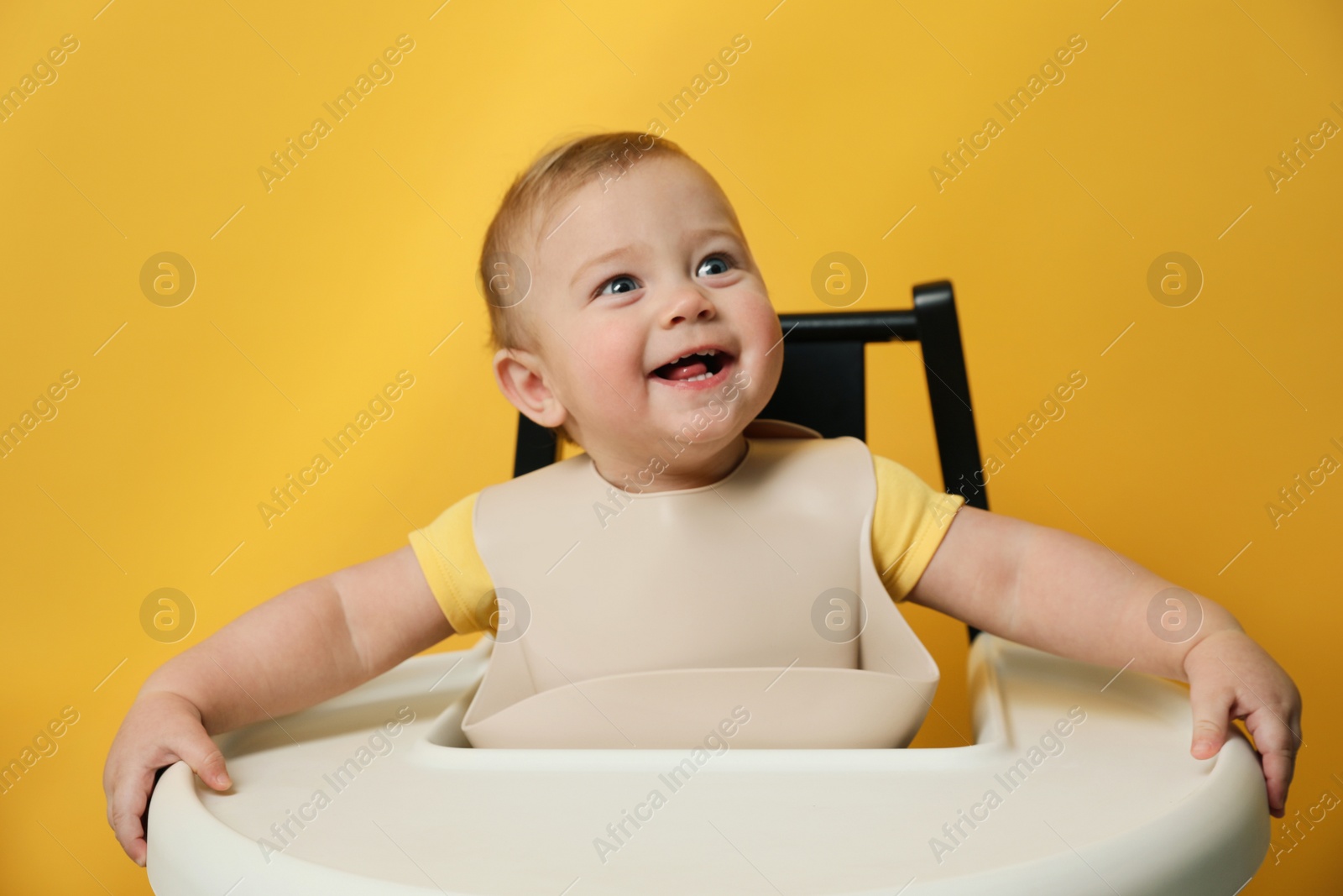 Photo of Cute little baby wearing bib in highchair on yellow background