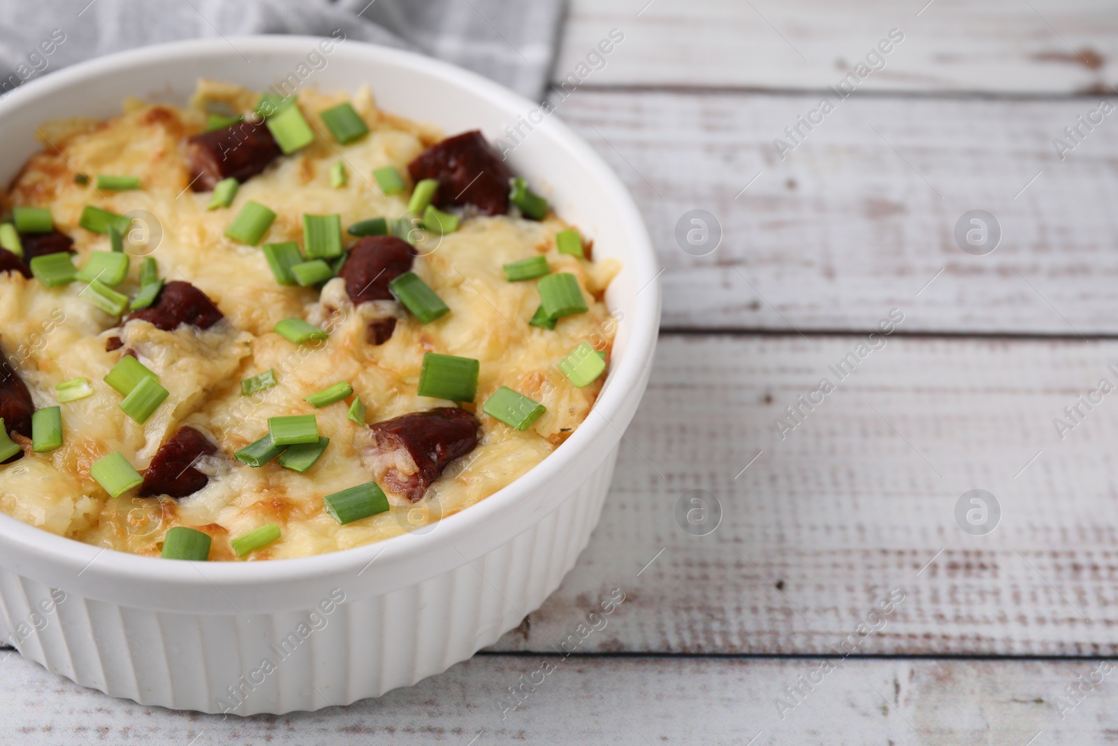 Photo of Tasty sausage casserole with green onion in baking dish on white wooden table, closeup. Space for text