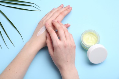 Woman applying hand cream on light blue background, top view