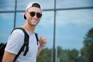 Photo of Handsome young man with stylish sunglasses and backpack near reflection surface outdoors, space for text