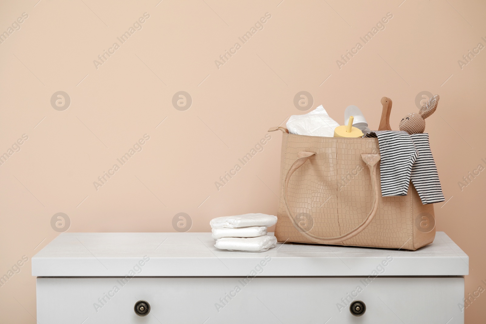 Photo of Mother's bag with baby's stuff on white chest of drawers near beige wall. Space for text