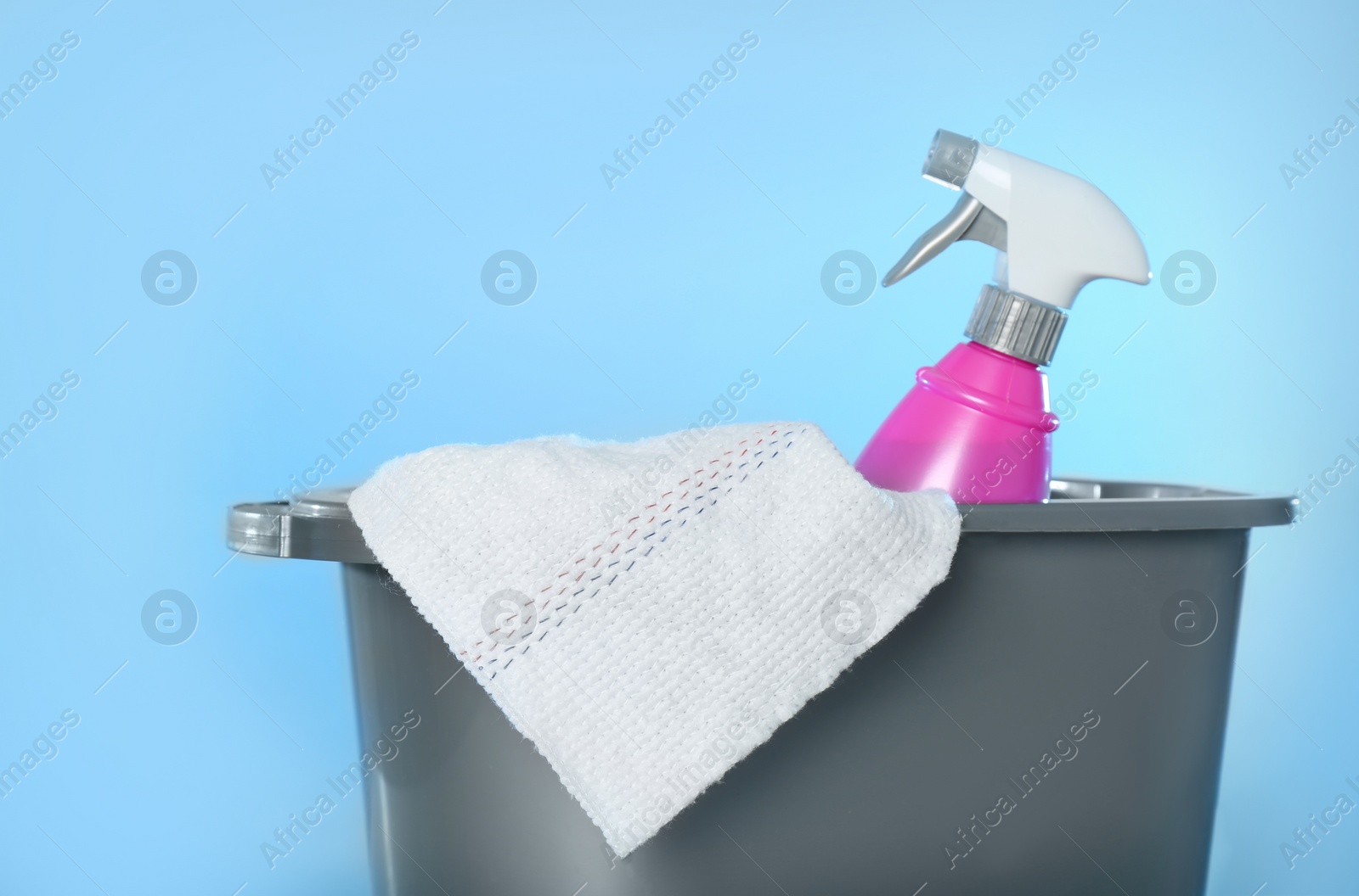 Photo of Bucket with bottle of cleaning product and microfiber cloth on light blue background, closeup