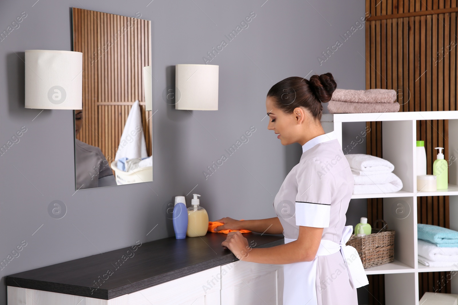 Photo of Young chambermaid wiping dust from furniture with rag in bathroom