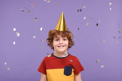Photo of Happy little boy in party hat under falling confetti on purple background