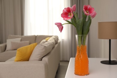 Photo of Orange filler with tulips in glass vase on white table at home, space for text. Water beads