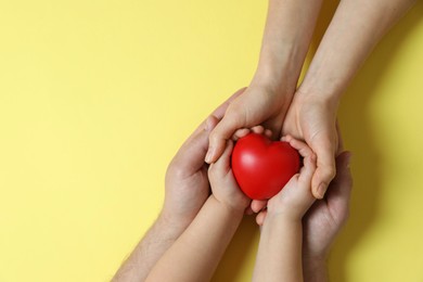 Parents and child holding red decorative heart on pale yellow background, top view. Space for text