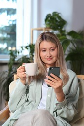 Photo of Woman with smartphone and cup of drink sitting in armchair at home