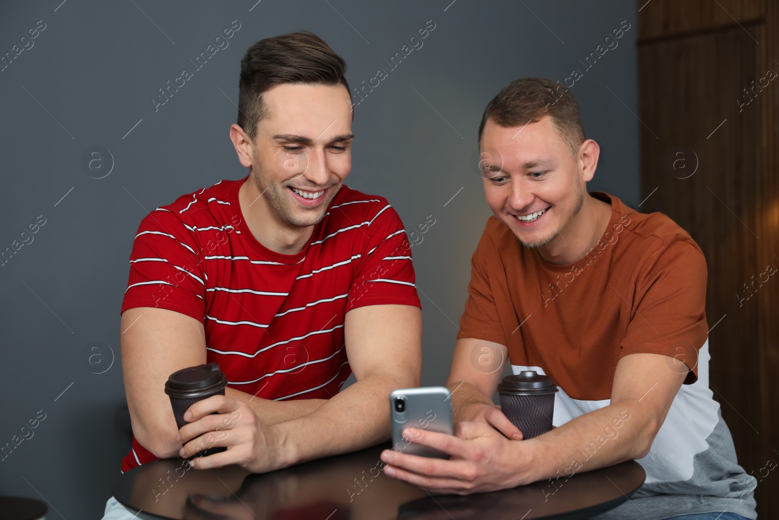 Photo of Men with cups of coffee and smartphone laughing together at table indoors