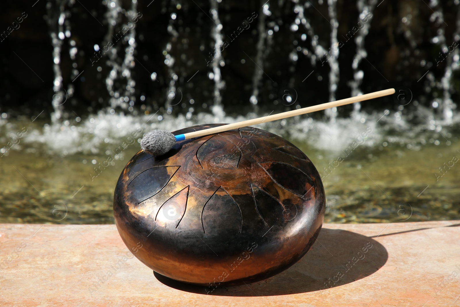 Photo of Steel tongue drum with mallet near waterfall outdoors on sunny day. Percussion musical instrument