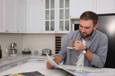 Photo of Handsome man with cup of coffee reading magazine at white marble table in kitchen