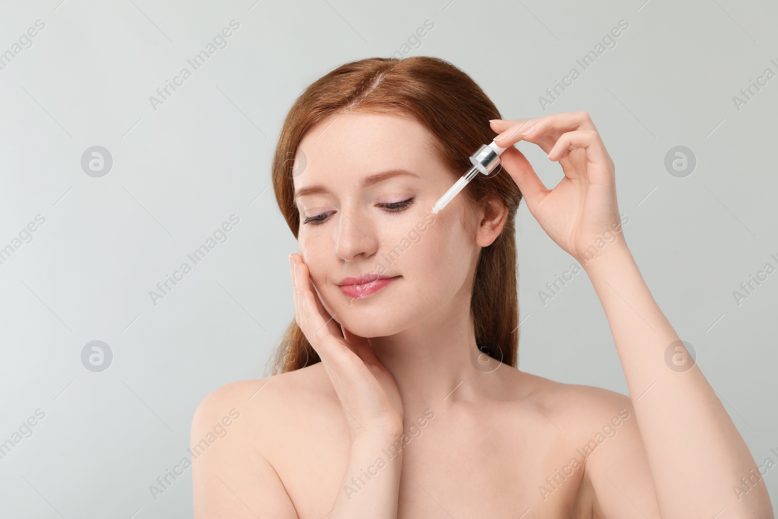 Photo of Beautiful woman with freckles applying cosmetic serum onto her face on grey background