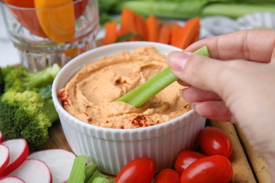 Photo of Woman dipping celery stick into hummus at table, closeup