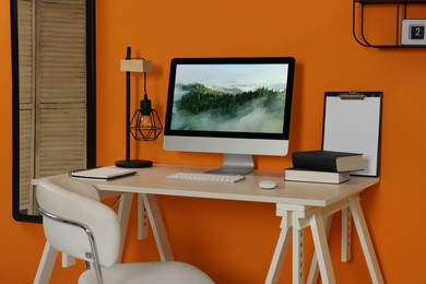Photo of Workplace with modern computer on wooden desk and comfortable chair near orange wall. Home office