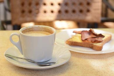 Photo of Cup of coffee and delicious sandwich with fried bacon on beige table