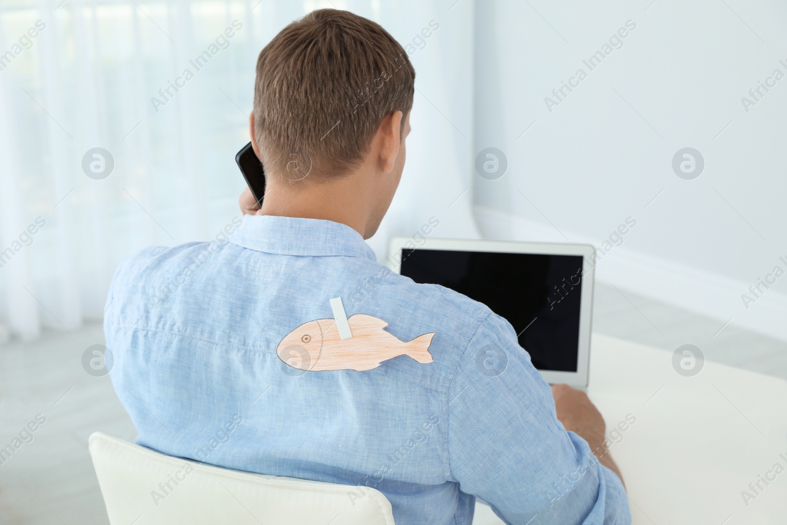 Photo of Man with paper fish on his back working with laptop at table indoors. April fool's day prank