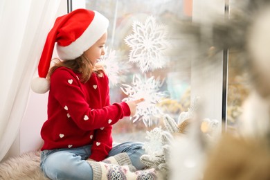 Photo of Little girl in Santa hat near window decorated with paper snowflakes at home