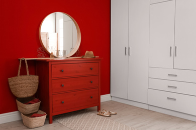 Round mirror and chest of drawers near red wall in room. Modern interior design