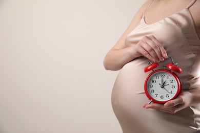 Young pregnant woman holding alarm clock near her belly on beige background, closeup and space for text. Time to give birth