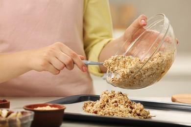Photo of Making granola. Woman putting mixture of oat flakes, dried fruits and other ingredients onto baking tray at table, closeup