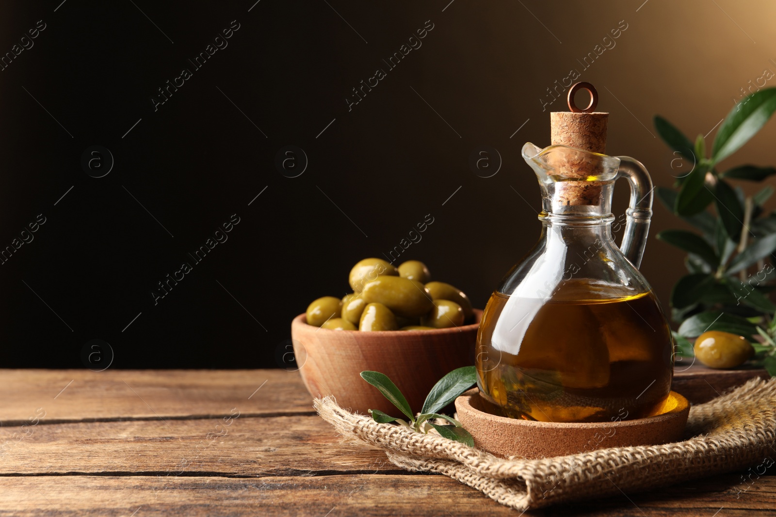 Photo of Glass jug of oil, ripe olives and green leaves on wooden table. Space for text