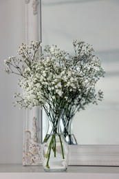 Beautiful gypsophila flowers in glass vase on white table near mirror indoors