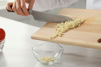 Professional chef putting cut garlic into bowl at white marble table indoors, closeup