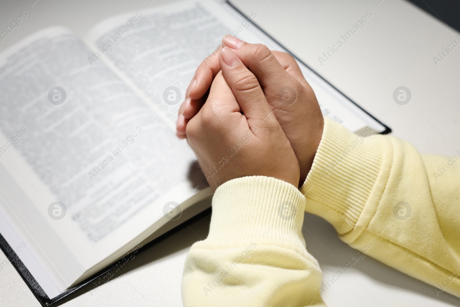 Photo of Woman holding hands clasped while praying over Bible at white table, closeup