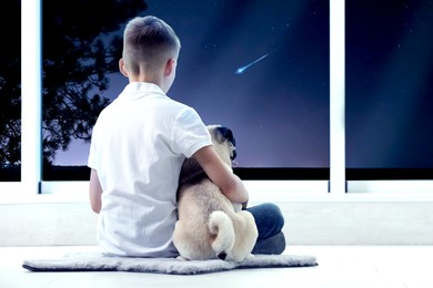 Cute little boy with dog sitting near window and looking at shooting star in beautiful night sky