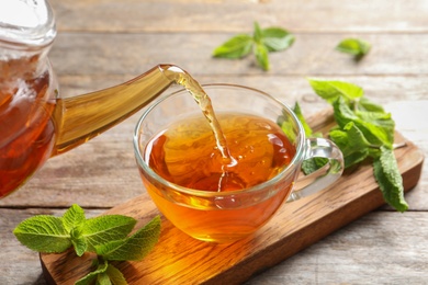 Photo of Pouring hot aromatic mint tea into cup on wooden board