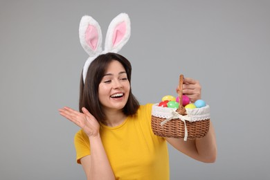 Easter celebration. Happy woman with bunny ears and wicker basket full of painted eggs on grey background