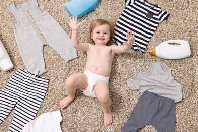 Photo of Little girl among baby clothes and detergents on carpet, top view