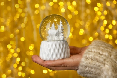 Woman holding snow globe with fir trees and deer against festive lights, closeup