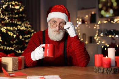 Photo of Santa Claus drinking hot beverage at his workplace in room decorated for Christmas