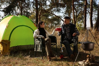 Photo of Couple sitting in camping chairs and clinking mugs outdoors