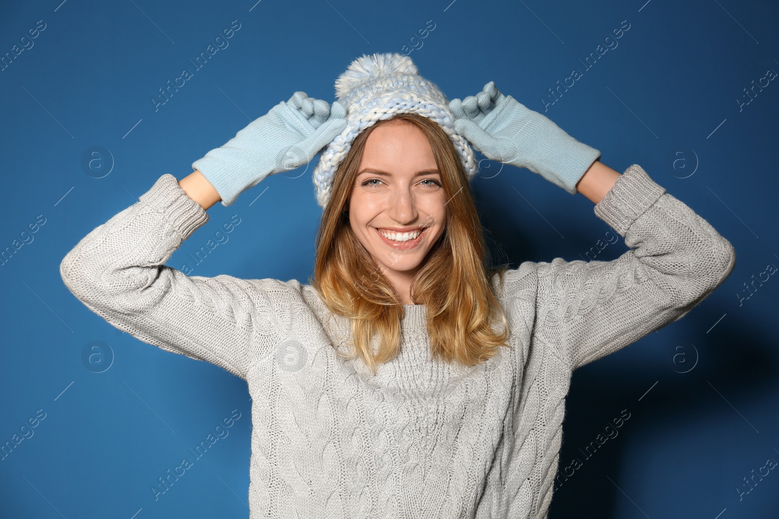 Image of Happy young woman wearing warm sweater, knitted hat and mittens on blue background