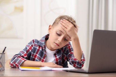 Photo of Little boy suffering from headache at wooden desk indoors